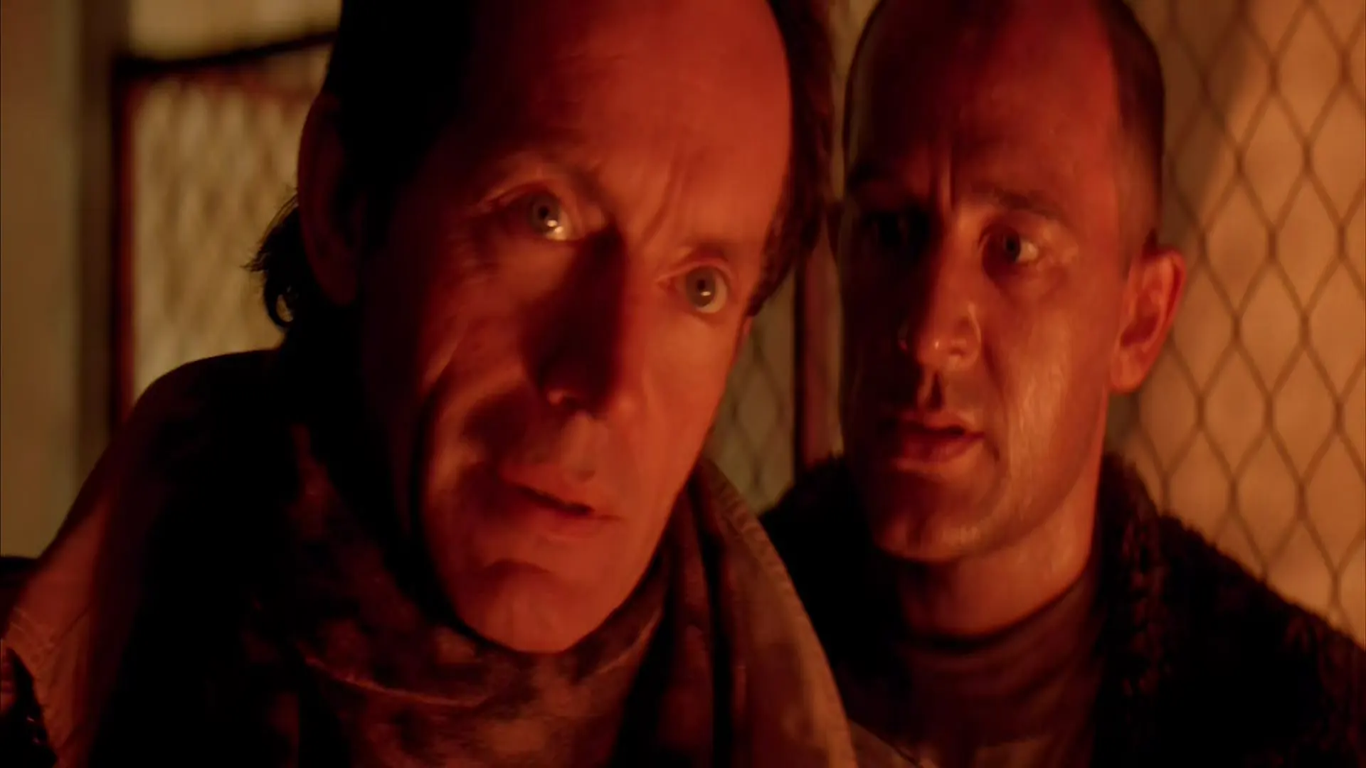 Company representative 'Bishop II' and another man confer in fiery light, their faces tense and illuminated by the furnace glow